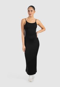 Zwarte maxi-jurk met spaghetti bandjes, een nauwsluitende pasvorm, een gladde textuur en een lengte tot de enkels, gecombineerd met witte sportschoenen.
