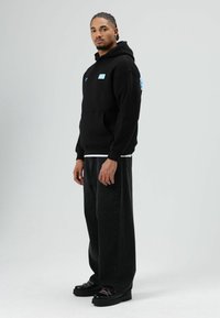 Sweat à capuche noir avec une poche avant et un logo bleu sur la poitrine gauche ; accompagné d'un jean noir ample et de chaussures noires à lacets.