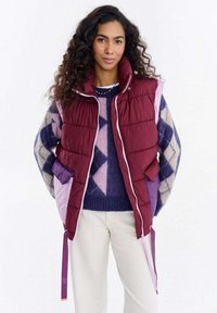 Bordowa pikowana kamizelka z fioletowymi akcentami i bocznymi kieszeniami, z wysokim kołnierzem i kontrastującymi zamkami błyskawicznymi, noszona na wzorzystym swetrze.
