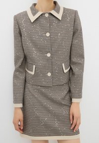 Brązowy i beżowy garnitur w kratę jodłową z kurtką z kołnierzykiem, pięcioma guzikami, dwiema kieszeniami i dopasowaną spódnicą. Teksturowany delikatnymi cekinami.