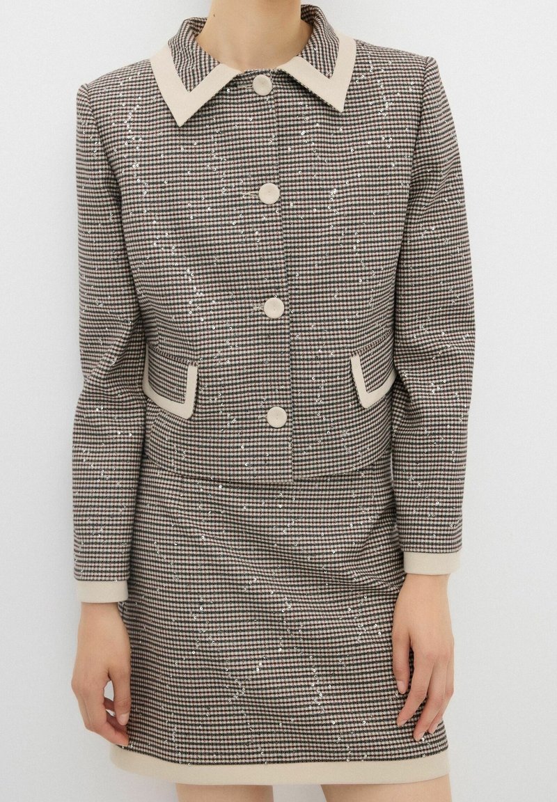 Brązowy i beżowy garnitur w kratę jodłową z kurtką z kołnierzykiem, pięcioma guzikami, dwiema kieszeniami i dopasowaną spódnicą. Teksturowany delikatnymi cekinami.