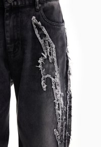 Pantalon en denim noir avec un grand patch vertical usé présentant des bords effilochés à côté de la poche extérieure de la cuisse.