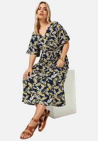 Marininspirerad blommig omlotttkjol med gula och vita bladmönster, kort ärm, knytmidja och en lång, flödande silhuett, i kombination med bruna sandaler.