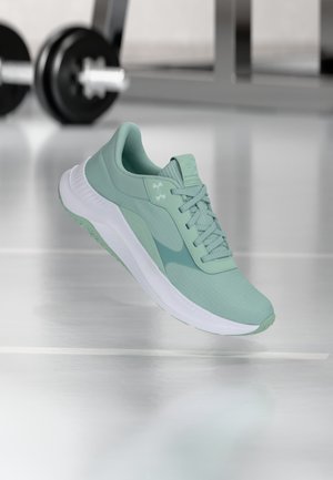 Hellgrüner Sportschuh mit weißer Sohle, der über einem reflektierenden Fitnessstudio-Boden schwebt, im Hintergrund unscharfe Gewichte.