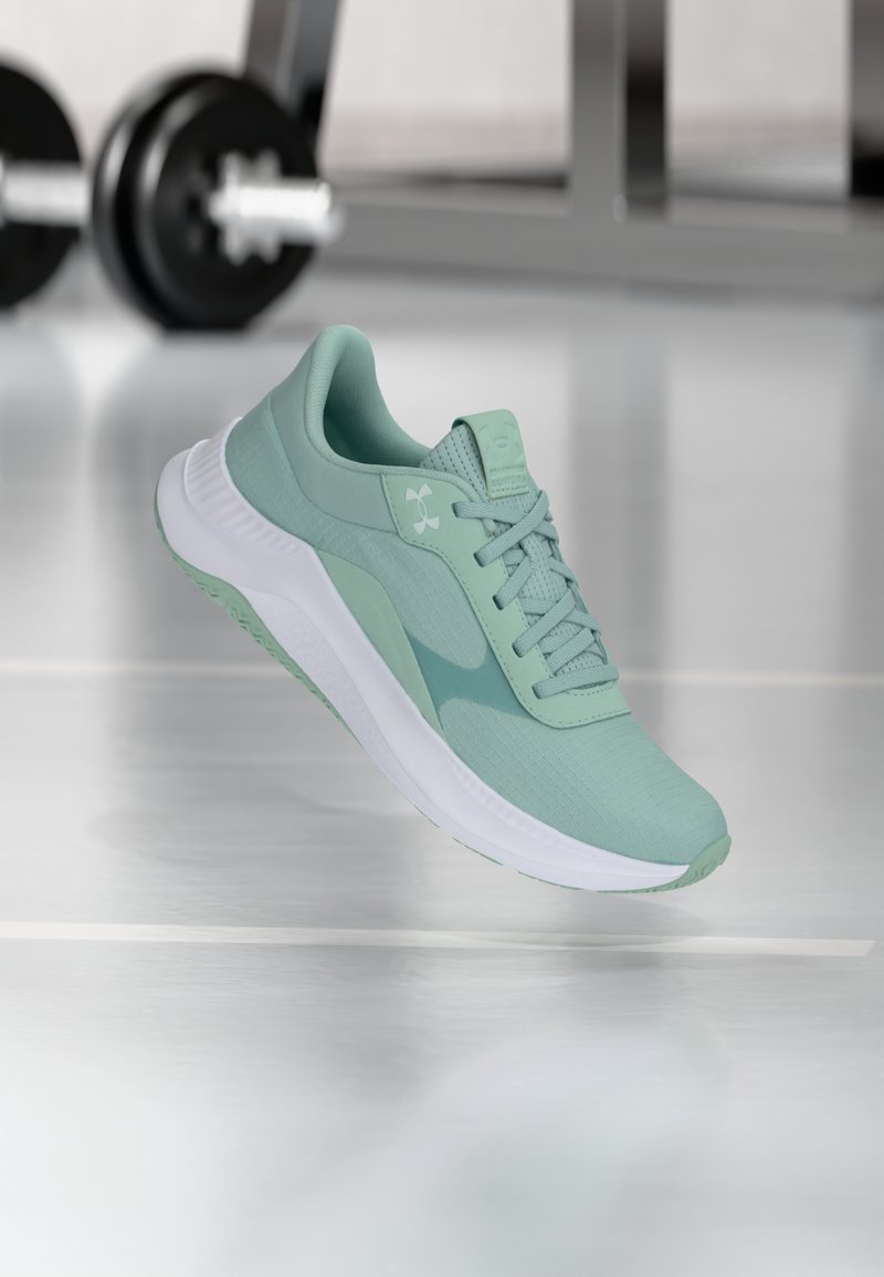 Αθλητικό παπούτσι σε ανοιχτό πράσινο χρώμα με λευκή σόλα, αιωρείται πάνω από αντανακλαστικό δάπεδο γυμναστηρίου, θολά βάρη στο φόντο.