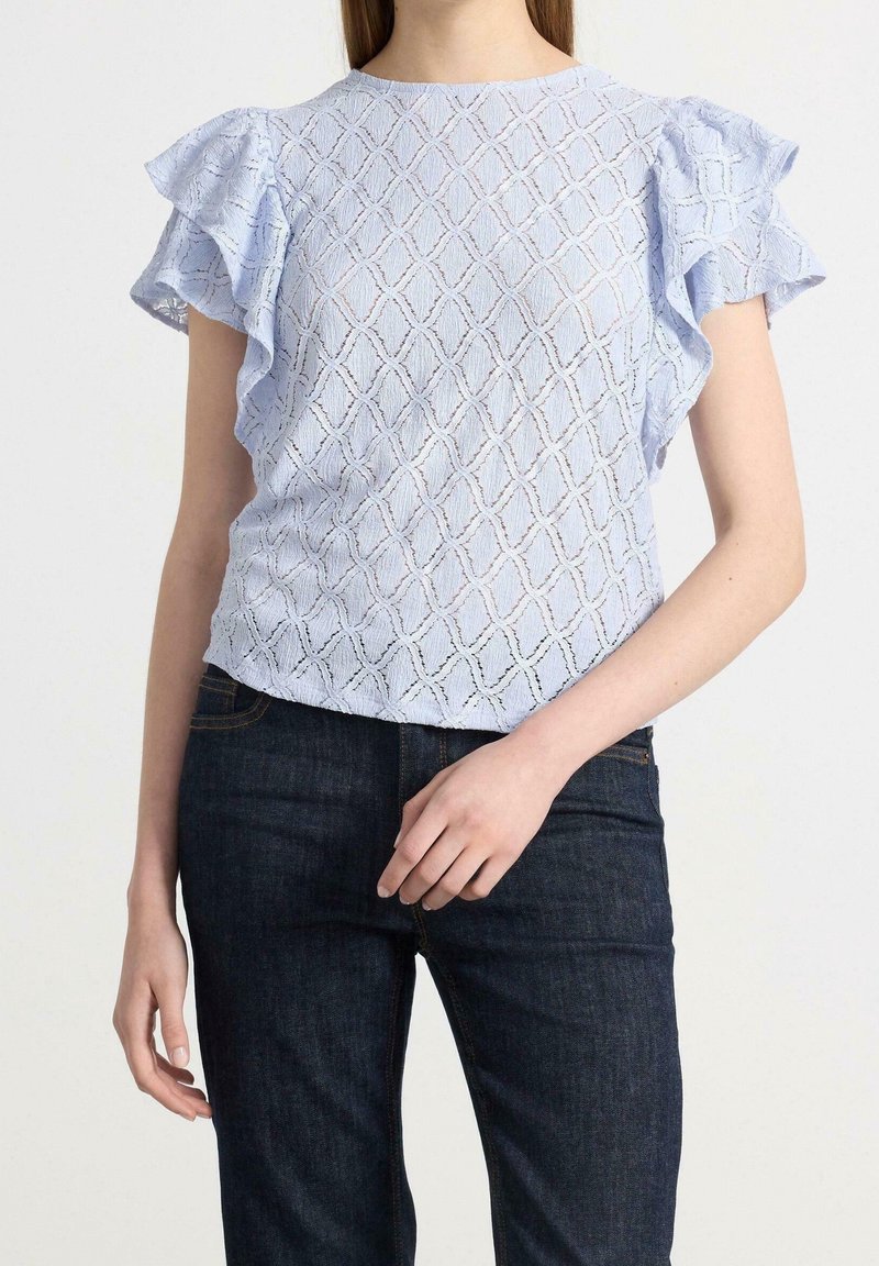 Femme portant un chemisier bleu clair à motifs avec des manches à volants et un jean bleu foncé, debout devant un fond blanc.