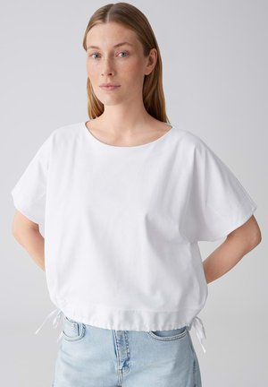 Giovane donna con capelli lisci castano chiaro che indossa una maglietta bianca larga a maniche corte e jeans azzurri, in piedi con le mani dietro la schiena.