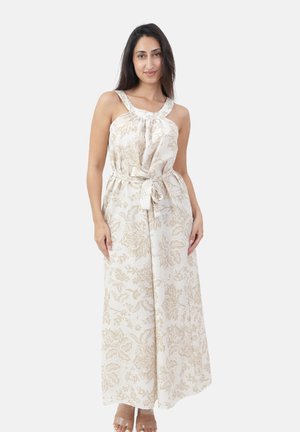 Vrouw in een mouwloze witte jumpsuit met een ceintuur en beige bloemmotieven, met doorzichtige sandaalhakken met bandjes.