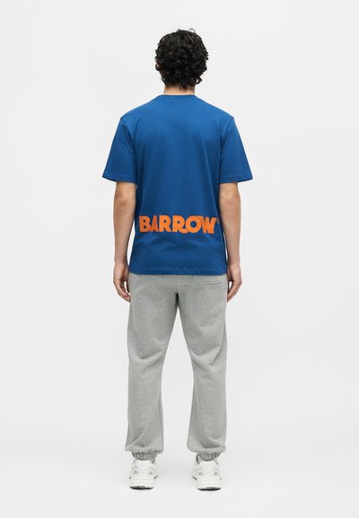 Άνδρας με σγουρά μαλλιά που φοράει μπλε μπλουζάκι με πορτοκαλί κείμενο "BARROW" στο κάτω μέρος της πλάτης, γκρι φόρμες και λευκά αθλητικά παπούτσια, όρθιος με την πλάτη προς το μέρος μας.