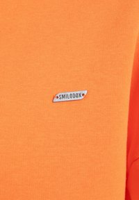Camiseta de algodón naranja con una pequeña etiqueta plateada que lleva el texto "SMILODOX" adjunta. Textura suave con un diseño ajustado.