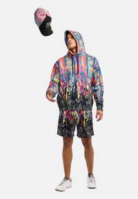 Bunte Kapuzenjacke mit abstraktem Wirbelmuster, passenden Shorts und einer schwarzen Mütze. Die Materialien wirken weich und die Jacke verfügt über eine verstellbare Kapuze.