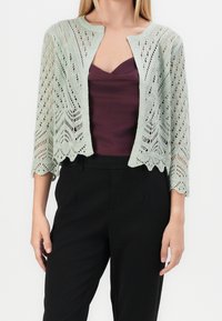 Světle zelený pletený cardigan s otevřeným designem, vlnitými okraji a rukávy po třičtvrtě, nošený přes tmavě fialový top a černé kalhoty.