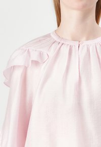 Blusa rosa realizzata in tessuto leggero, con maniche arricciate, scollatura rotonda con dettagli arricciati e chiusura a bottone sul retro.