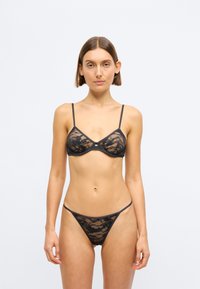 Crni čipkani donji veš uključuje bralet s tankim trakama i odgovarajući tange, oboje s cvjetnim uzorkom i prozirnom teksturom.