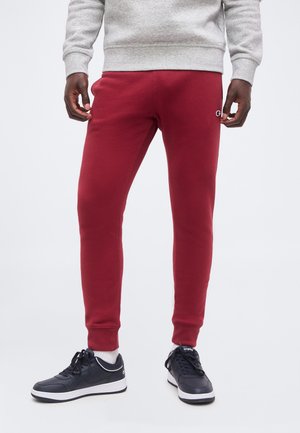 Mann trägt rote Slim-Fit-Jogginghosen, graues Sweatshirt, weiße Socken und schwarze Sneakers und steht vor einem weißen Hintergrund.