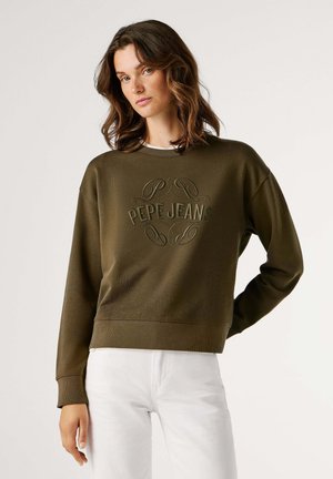 Femme aux cheveux ondulés portant un sweat-shirt vert olive avec le logo "PEPE JEANS" et un pantalon blanc, posant devant un fond neutre.