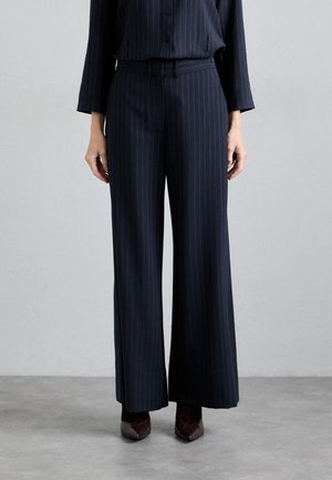 Pantalons larges à rayures noires avec une texture lisse, dotés d'une taille ajustée et de poches latérales discrètes, associés à des chaussures pointues foncées.