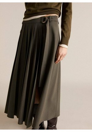Frau trägt einen plissierten olivgrünen Midirock mit seitlichem Gürteldetail, kombiniert mit einem braunen Langarmshirt und dunklen Stiefeln.