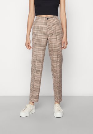Pantalon beige à carreaux slim porté avec des baskets blanches à plateforme et un top court noir, debout sur un sol gris avec un fond blanc.