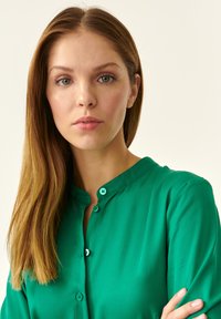 Blusa verde con un cierre de botones en la parte delantera y una textura suave. Presenta un cuello puntiagudo y mangas largas. La tela parece ligera y suave.
