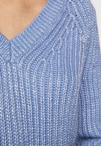 Světlomodrý pletený svetr s V výstřihem, představující žebrovanou texturu a svislé čáry. Příze obsahuje jemné třpytivé akcenty.