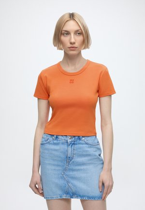 Kvinde med kort blondt hår iført en orange tætsiddende T-shirt og en lyseblå denim nederdel står mod en ensfarvet hvid baggrund.