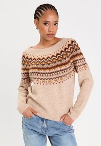 Woman with braided hair wearing beige sweater with multi-color zigzag pattern and blue jeans, posing with hands in pockets.