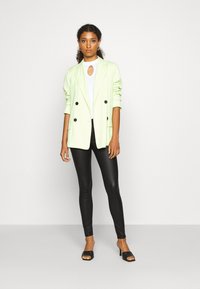 Blazer vert clair à double boutonnage avec des boutons noirs, porté sur un haut blanc et associé à des leggings noirs brillants et des talons ouverts noirs.