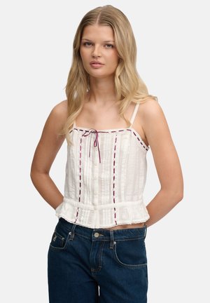 Ung kvinde med langt blondt hår iført hvid ærmeløs bluse med lilla syninger og blå jeans, stående med hænderne bag ryggen.