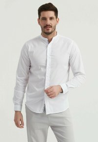 Chemise blanche à boutons avec col montant, manches longues et ourlet légèrement arrondi. Présente des coutures décoratives à l'intérieur du col.