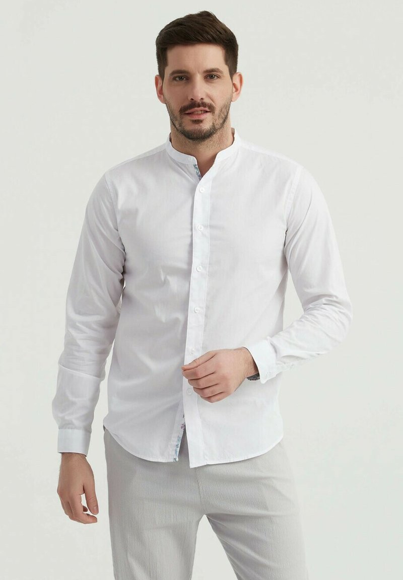 Chemise blanche à boutons avec col montant, manches longues et ourlet légèrement arrondi. Présente des coutures décoratives à l'intérieur du col.