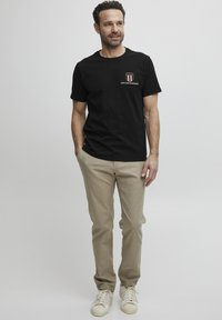 Juodos medvilnės marškinėliai su krūtinės logotipu; derinami su smėlinėmis kelnėmis ir baltomis sportbačiais. Tiesus fasonas, trumpomis rankovėmis ir apvaliu iškirpimu.