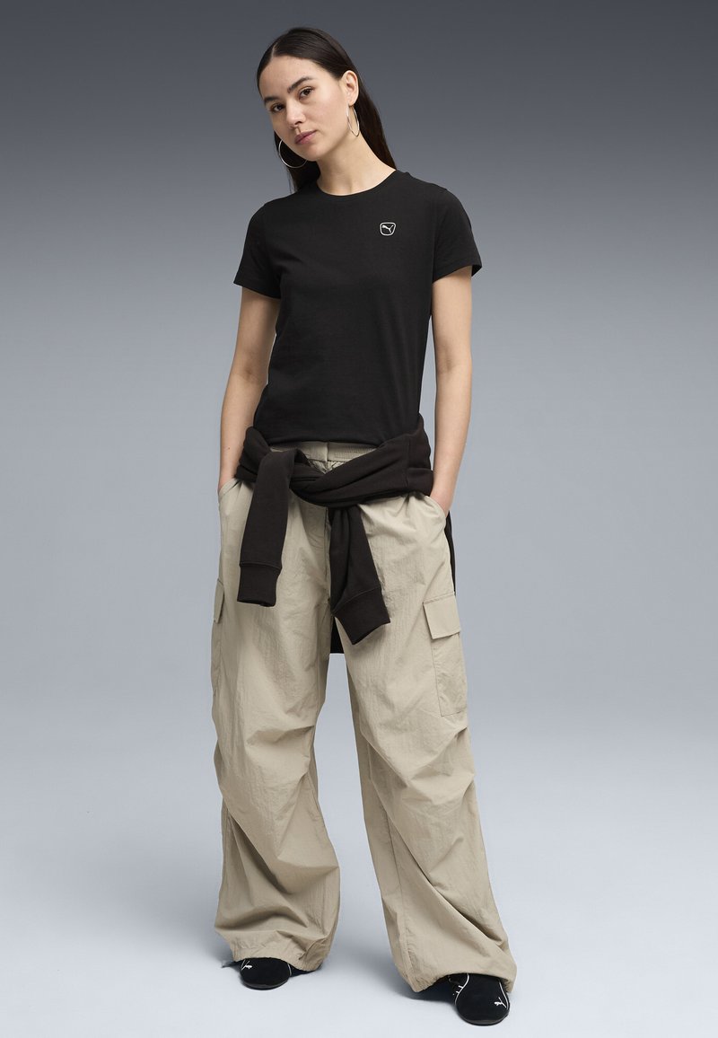 Femme debout les mains dans les poches, portant un t-shirt noir à manches courtes, un pantalon cargo beige à jambes larges, un sweat-shirt noir noué autour de la taille et des chaussures noires.