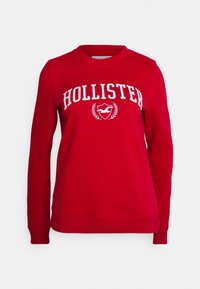 Sudadera roja con la palabra "HOLLISTER" en letras blancas, con un diseño de laurel y logo en el pecho. Cuello redondo simple y puños acanalados.