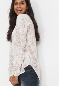 Blusa floreale con sfondo bianco, fiori rosa e gialli, maniche lunghe, vestibilità comoda e spacchi laterali all'orlo.