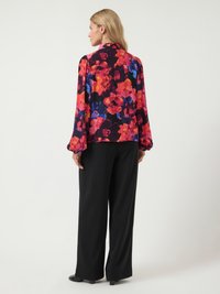 Mulher de costas, vestindo uma blusa de manga longa com padrões florais em vermelho, laranja e azul, e calças largas pretas.