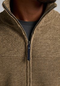 Chaqueta de punto con cremallera en beige, de tela texturizada y cuello de canalé. Presenta una cremallera plateada y una camiseta oscura visible debajo.