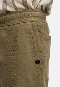 Pantalones de algodón color caqui con tejido texturizado, que cuentan con bolsillos laterales y una etiqueta negra de marca en la costura. Diseño estructurado con un ajuste entallado.