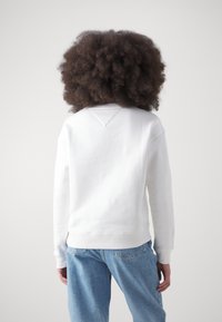 Persona con capelli ricci che indossa un semplice sudario bianco e jeans blu, in piedi con le spalle rivolte verso l'osservatore su uno sfondo neutro.