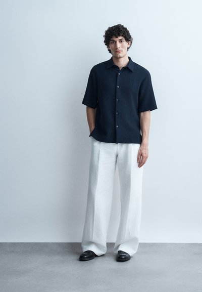 Άνδρας με σγουρά μαλλιά που φοράει μαύρο πουκάμισο με κοντά μανίκια και κουμπιά, λευκό παντελόνι φαρδύ στο πόδι και μαύρα παπούτσια, στέκεται μπροστά σε απλή τοίχο.