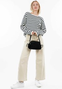 Borsa a tracolla nera in tessuto con manico intrecciato, abbinata a una camicia a righe blu navy e bianche a maniche lunghe e pantaloni beige a gamba larga. Sneakers bianche.