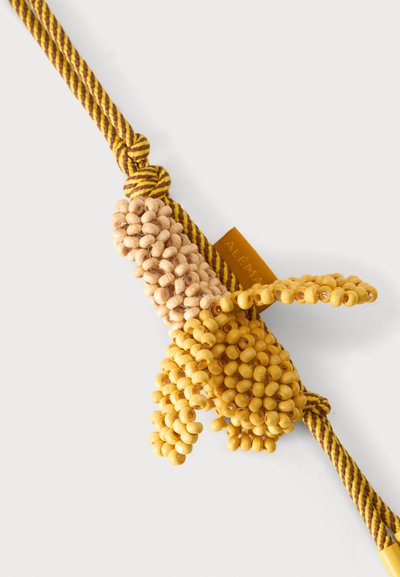 Gland jaune et beige orné de perles fixé à un cordon torsadé jaune et marron avec une petite étiquette en tissu portant l'inscription "ALEMA".