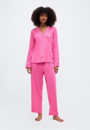 Žena v jasne ružovej dlhšie zapínanej pyžamovej blúzke a zhodných voľných nohaviciach stojaca bosá na bielom pozadí.