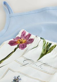 Débardeur côtelé bleu clair associé à un pantalon blanc à taille élastique avec des imprimés floraux botaniques colorés et du texte en écriture cursive.