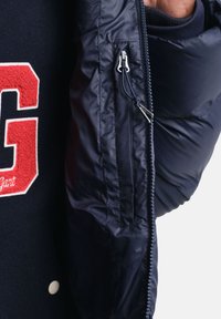 Marineblå pufferjakke med rød broderet bogstav "G" og cremefarvede knapper. Inkluderer en sølv lynlås og indvendig lomme med lynlåslukning.