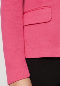 Ljust rosa texturerad tröja med rundad halsringning. Har en bröstficka och en figurnära silhuett, kombinerad med en svart underdel.