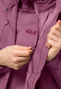 Des mains fermant la fermeture éclair d'une veste d'extérieur violette, dévoilant un vêtement intermédiaire violet avec le logo "Jack Wolfskin" sur la poitrine.