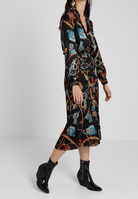Abito midi nero con catena colorata e stampa floreale, dotato di colletto e maniche lunghe, abbinato a stivaletti neri lucidi.