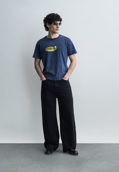 Άνδρας με σγουρά μαλλιά που φοράει γυαλιά ηλίου, μπλε γραφικό μπλουζάκι, φαρδιά μαύρη παντελόνα και μαύρα παπούτσια, στέκεται με τα χέρια στις τσέπες.