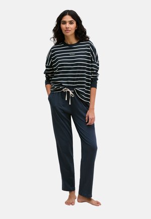 Vrouw staat blootsvoets, draagt een zwart-wit gestreept shirt met lange mouwen en donkerblauwe broek met trekkoord, tegen een effen achtergrond.